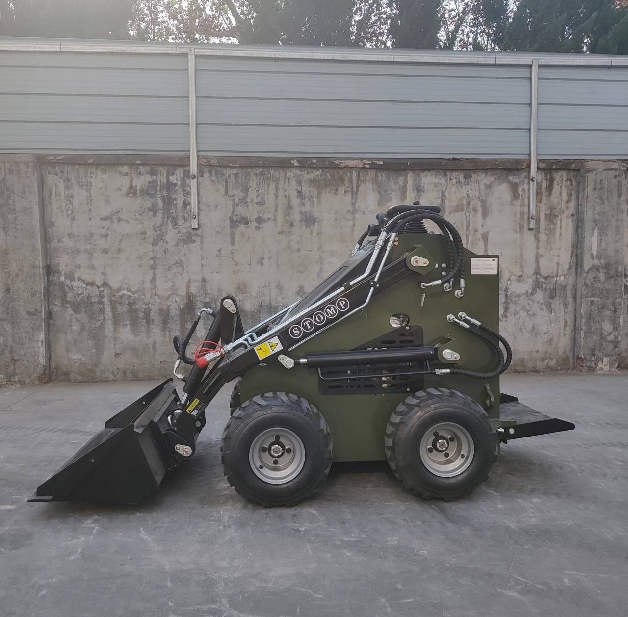 Compact olive-green skid-steer loader with a front bucket, parked against a concrete wall in an industrial yard