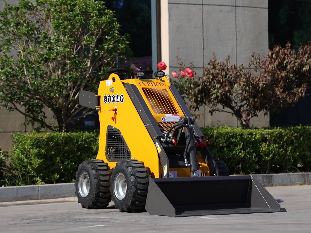 Yellow skid-steer loader with a wide front bucket parked on concrete, outdoors by bushes and a building.