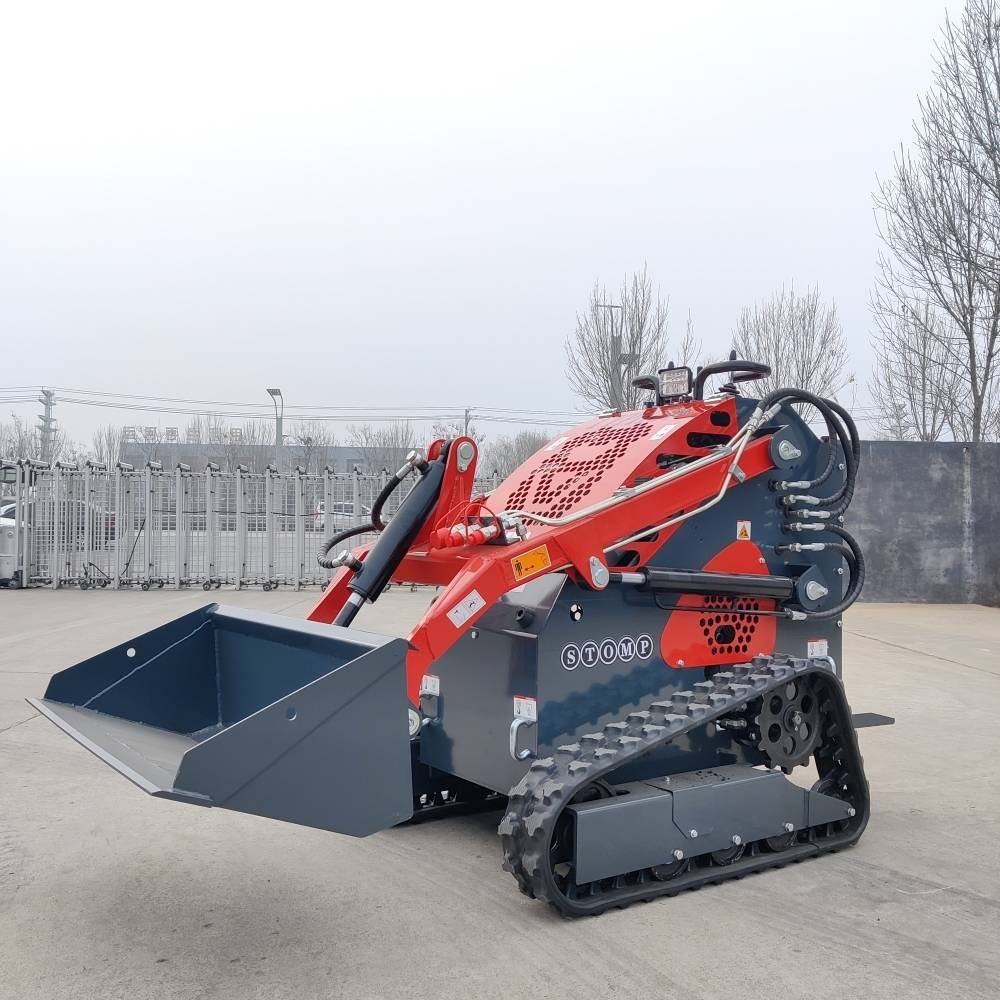 Red and gray compact tracked loader with front bucket, parked on concrete outdoors, industrial equipment rigging visible in background