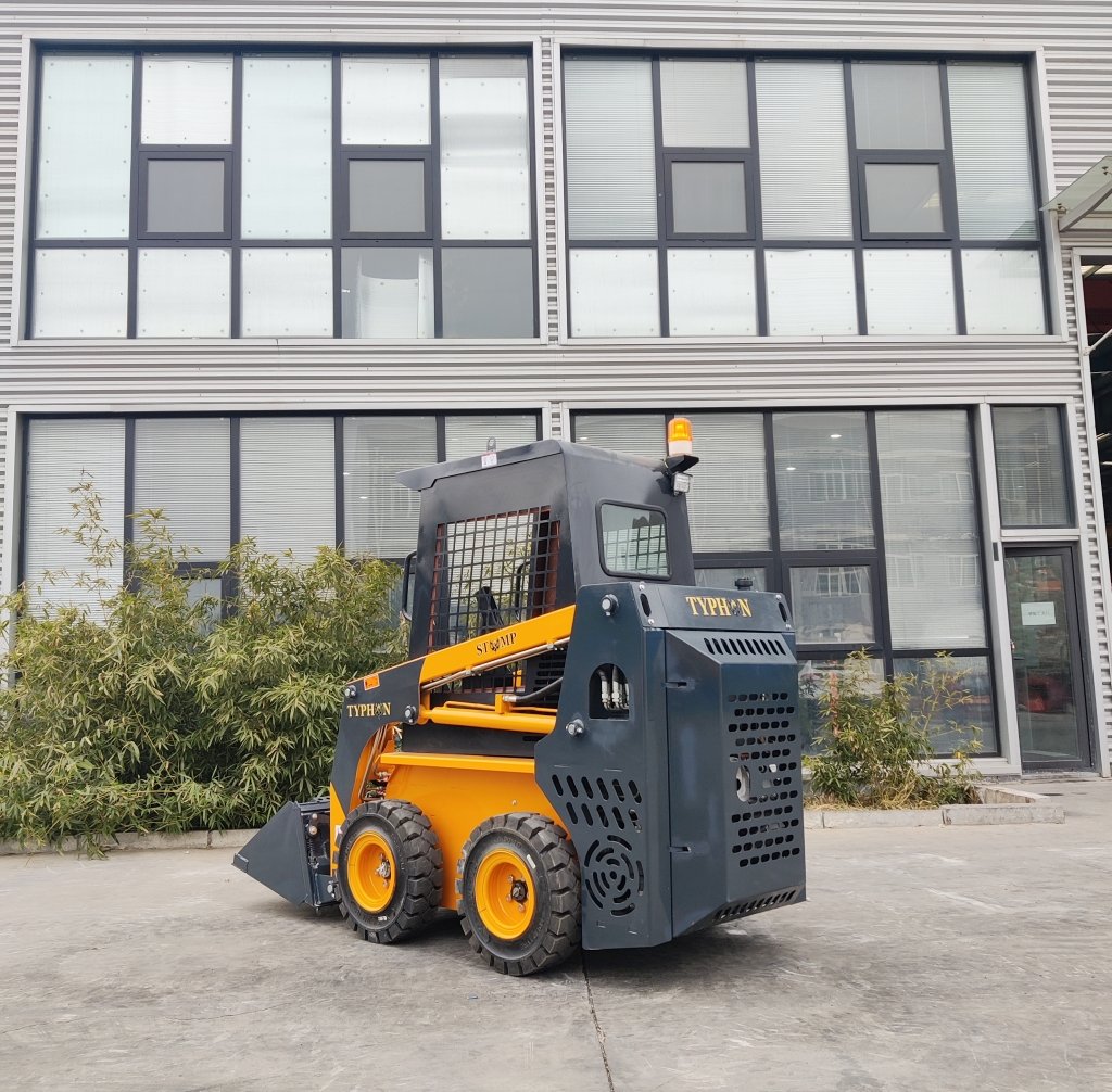 Small skid-steer loader with a front bucket, parked in front of a modern glass-walled building; yellow and dark blue colors with orange accents.