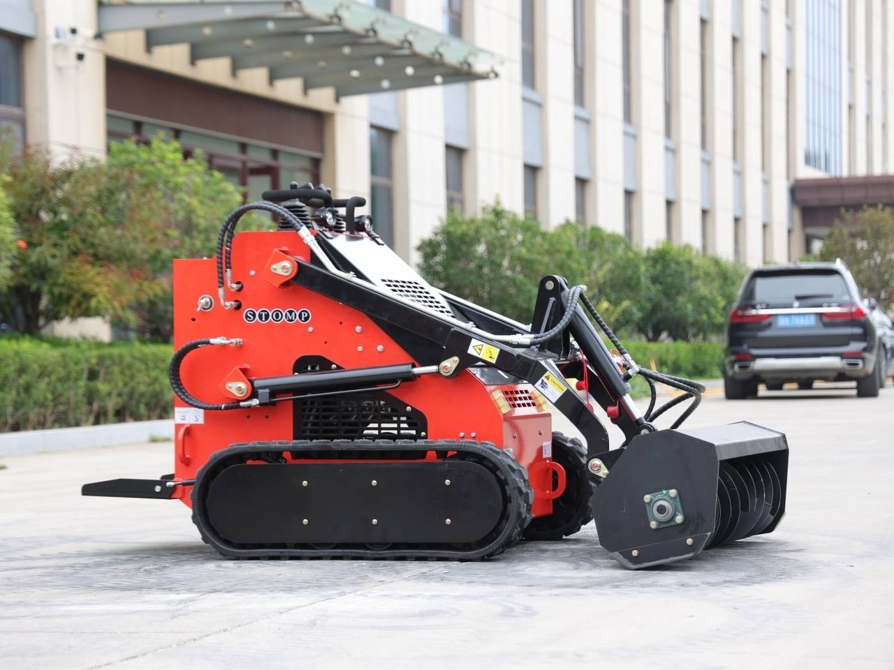 Red compact tracked loader with a large front bucket attachment, parked on a paved area in front of a building complex