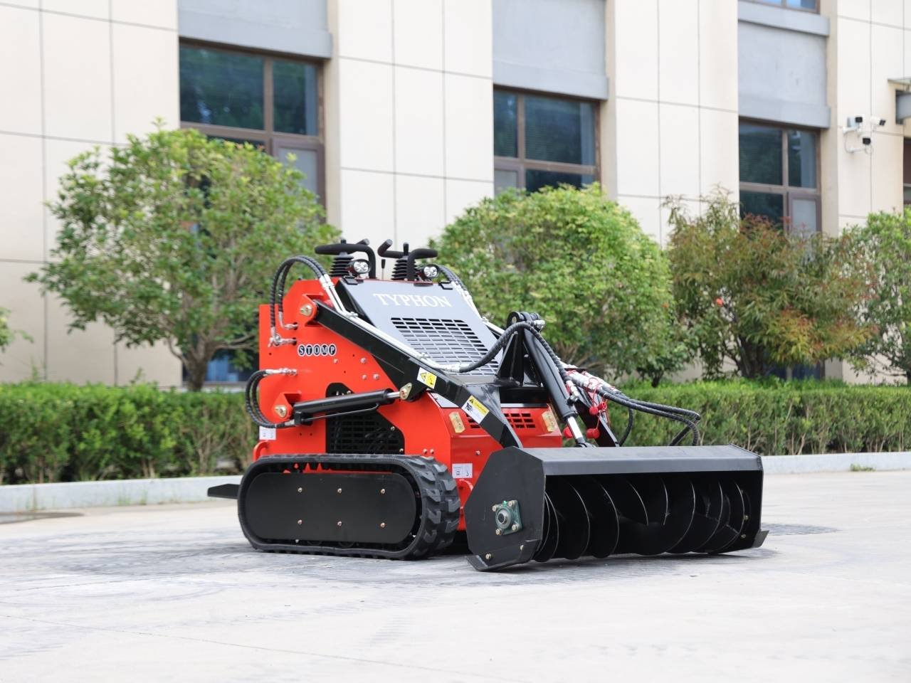 Red compact tracked machine with a large front-mounted auger attachment, labeled TYPHON, parked on a paved area in front of a beige building.
