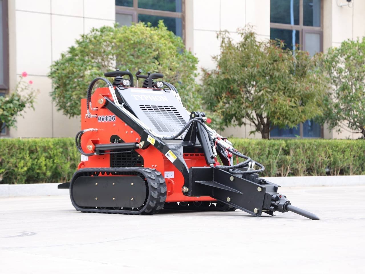 Red compact tracked loader with a hydraulic front attachment, parked on concrete near greenery; brand name Typhon visible on the grill.