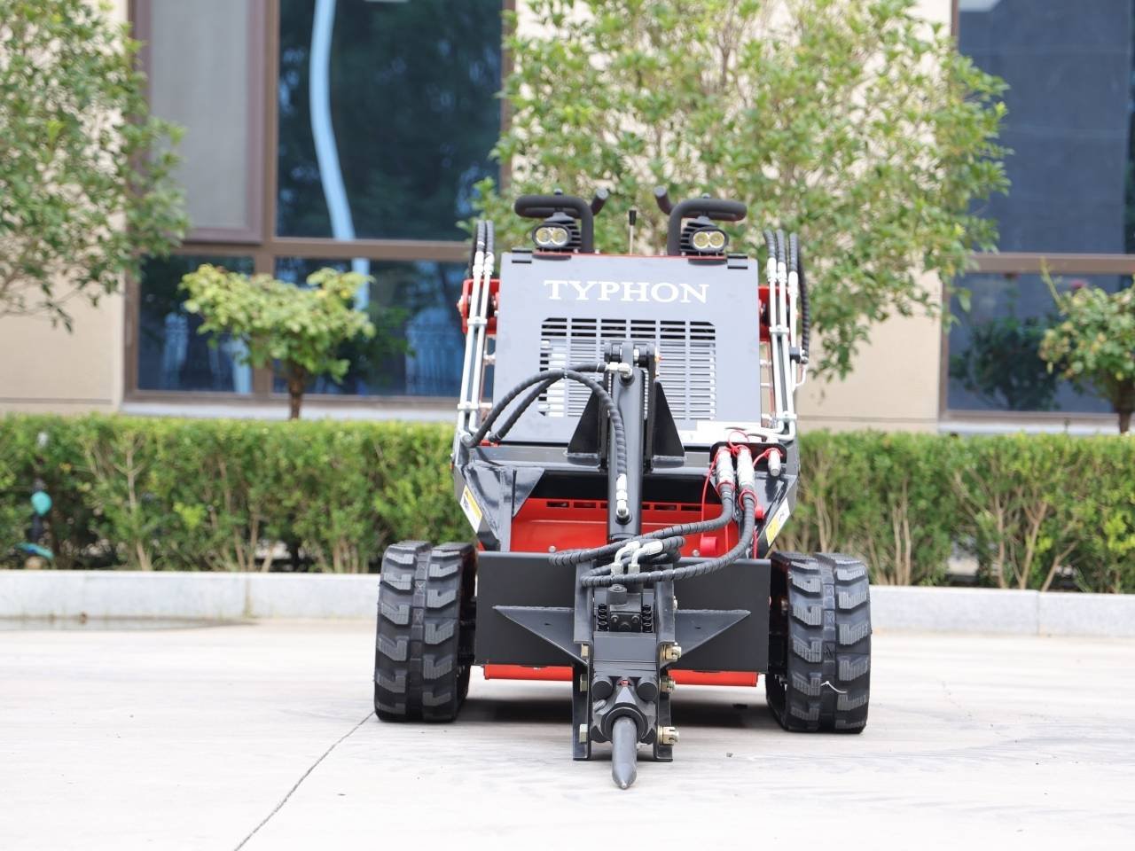 Front view of a gray and red Typhon industrial machine with large off-road tires and hydraulic arms on a concrete plaza, outdoors.