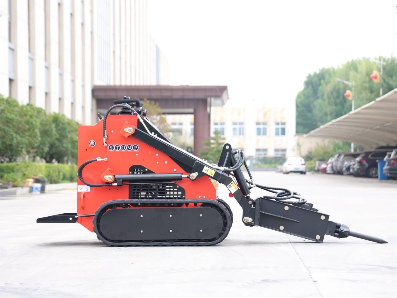 Red compact crawler excavator with a black hydraulic arm parked on a concrete surface near office buildings.