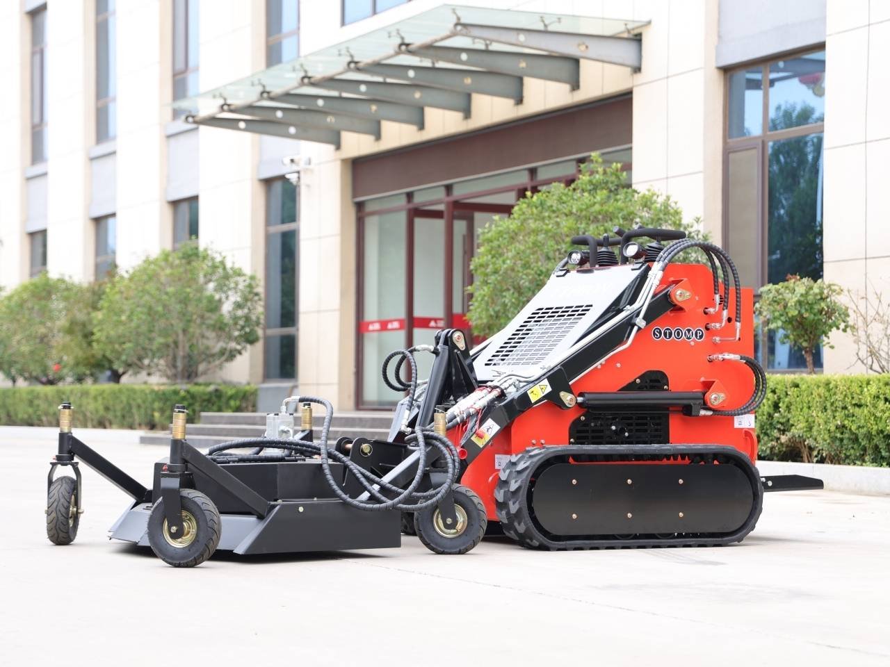 Red tracked mini loader with a large black front attachment parked on a concrete plaza in front of a modern office building.