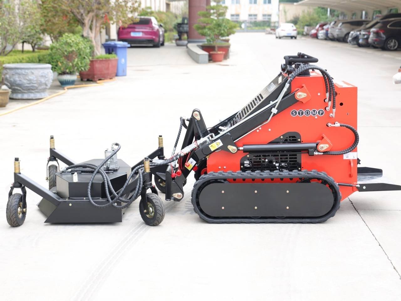 Red compact tracked machine with a detachable wheeled attachment in a parking lot, likely construction or landscaping equipment (STOMP brand).