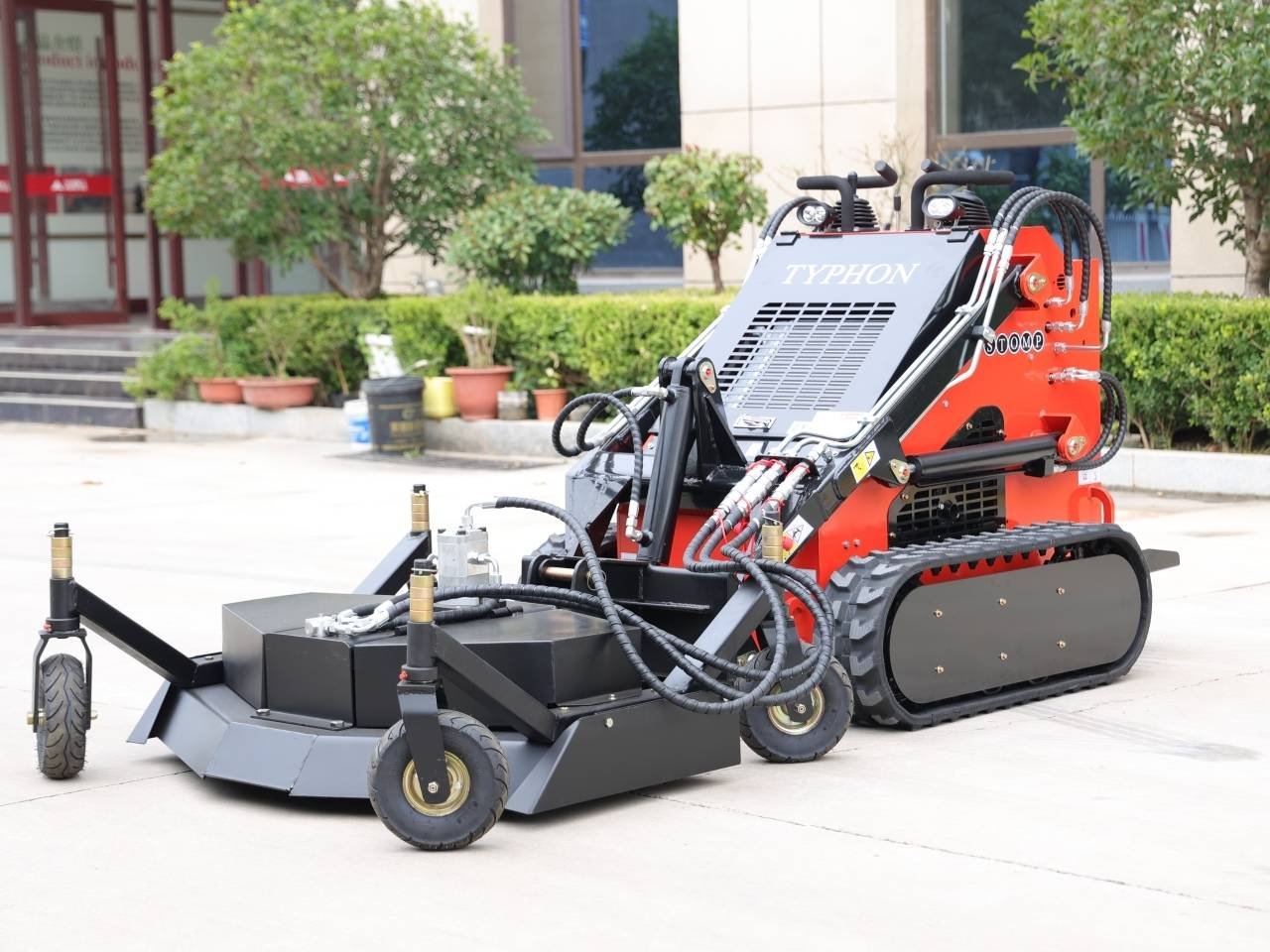 Red and black track-mounted road milling machine with hydraulic hoses parked on a concrete surface.