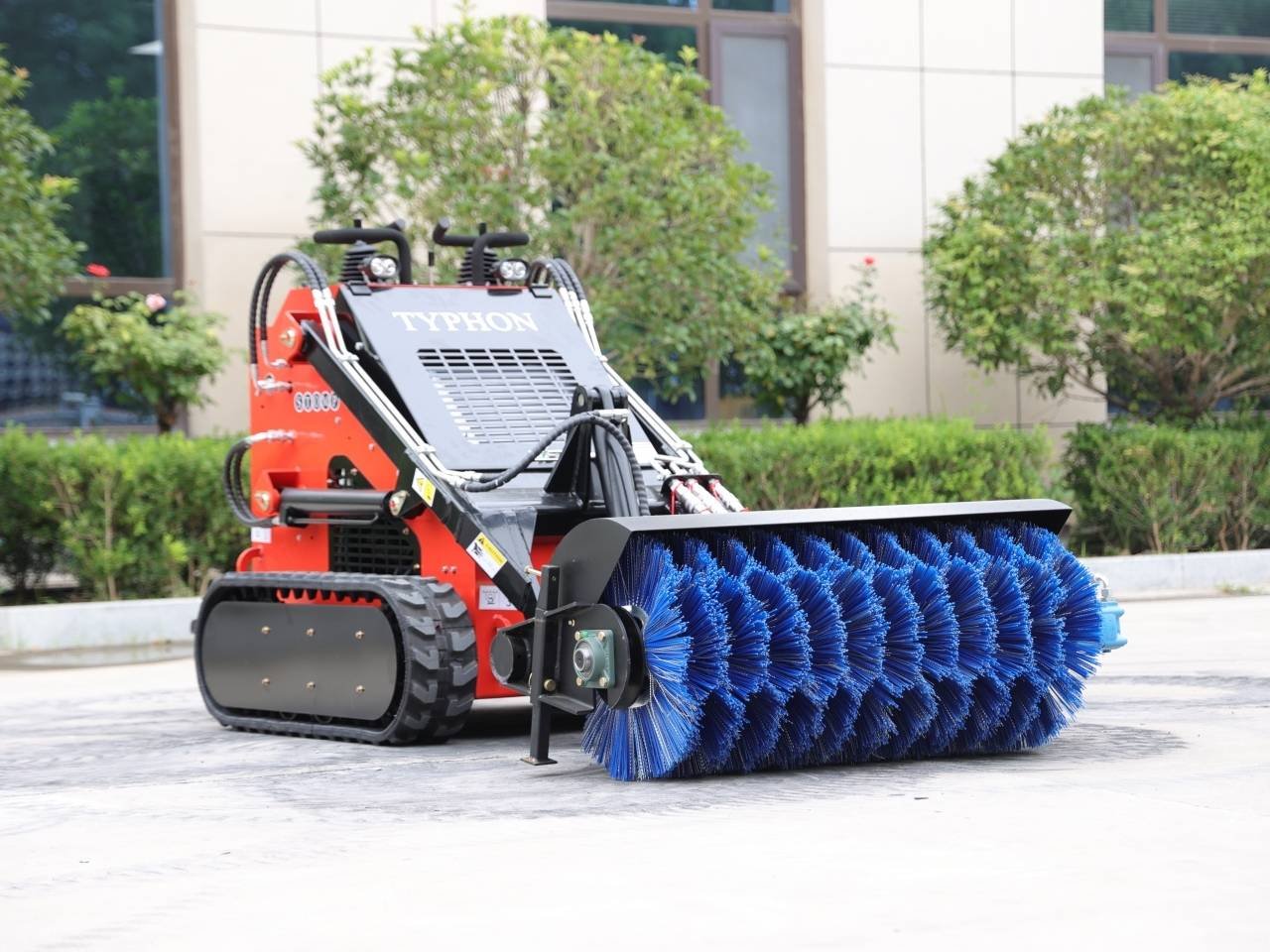 Red compact skid-steer sweeper with a large blue cylindrical brush on a paved area.