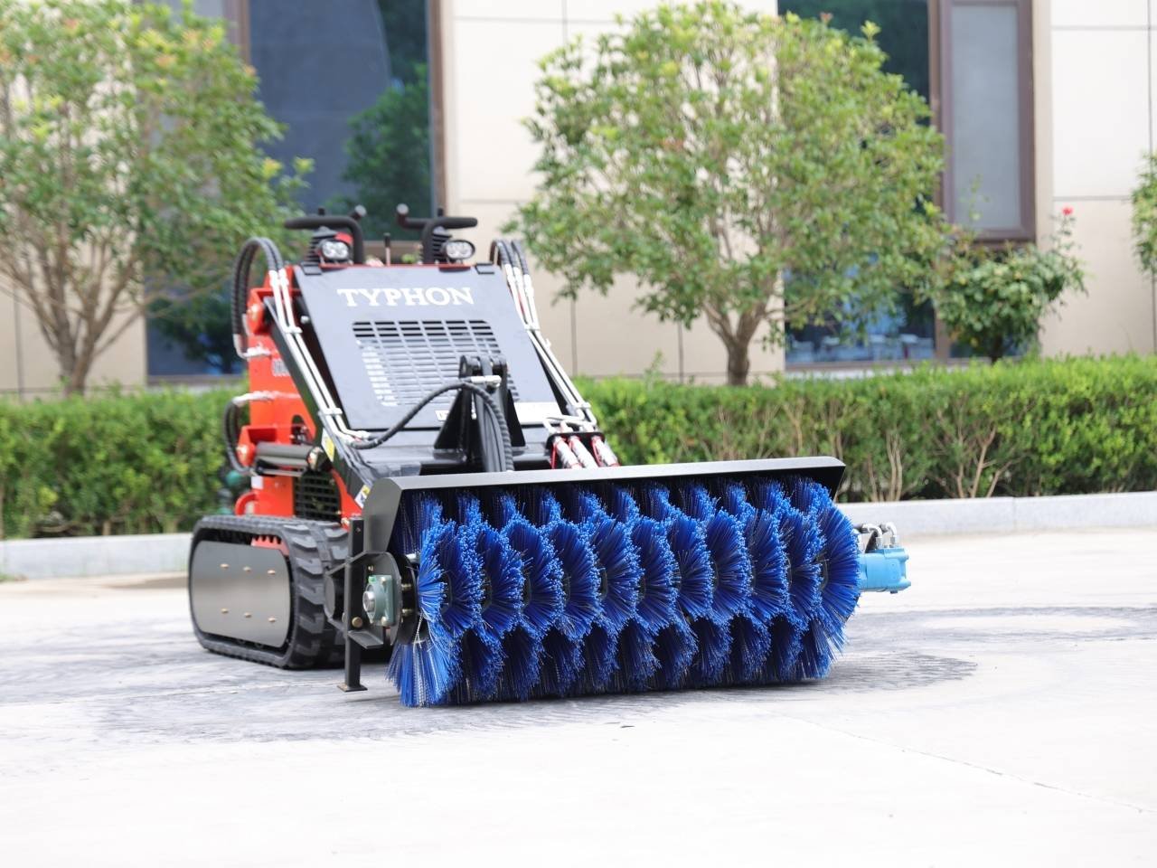 Red compact street sweeper on tracks with a large blue cylindrical brush in the front, parked on a paved area near greenery and a building, labeled TYPHON