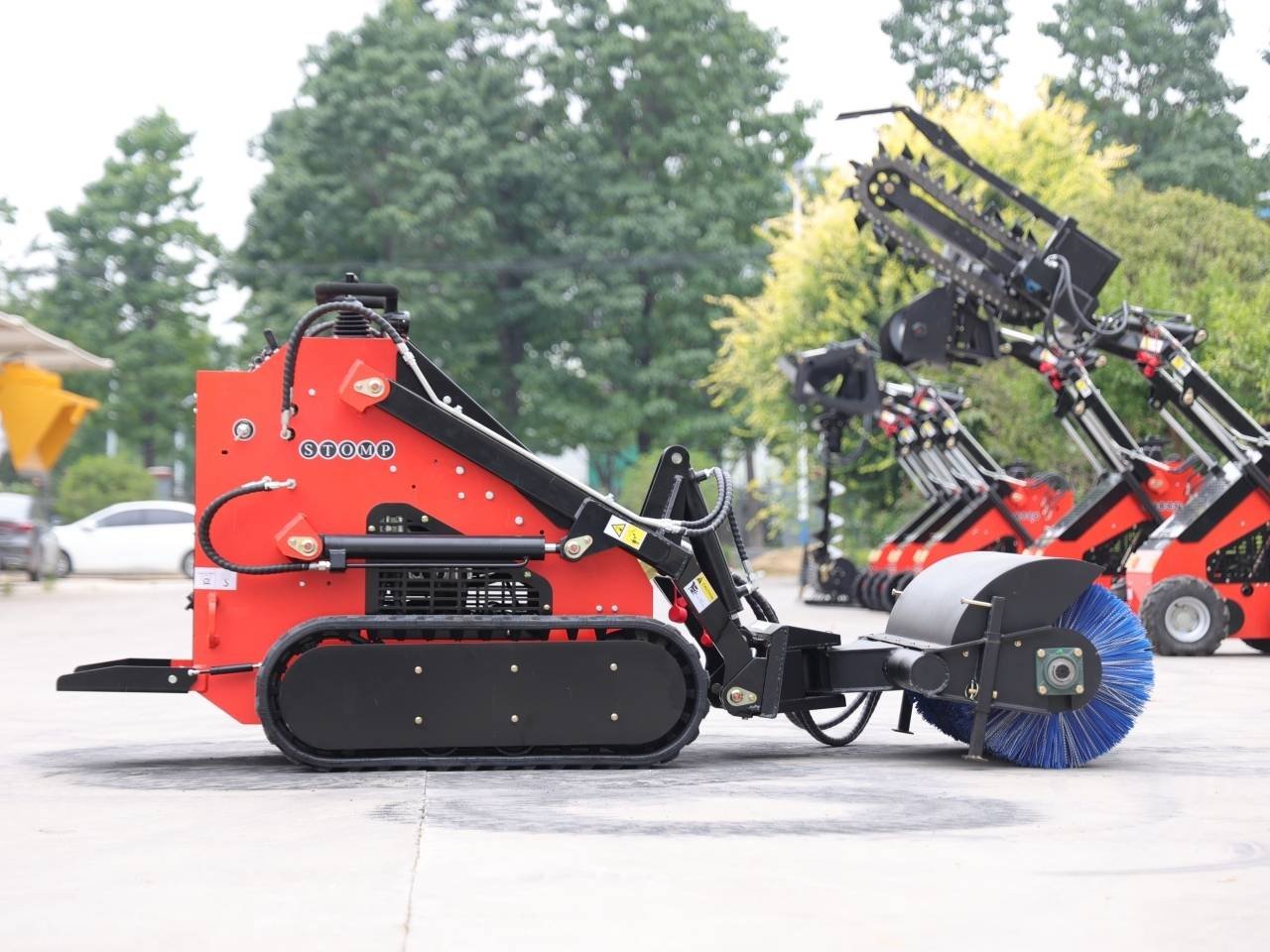 Red tracked street cleaning machine with a large blue rotating brush at the front, parked on a pavement with more red units in the background