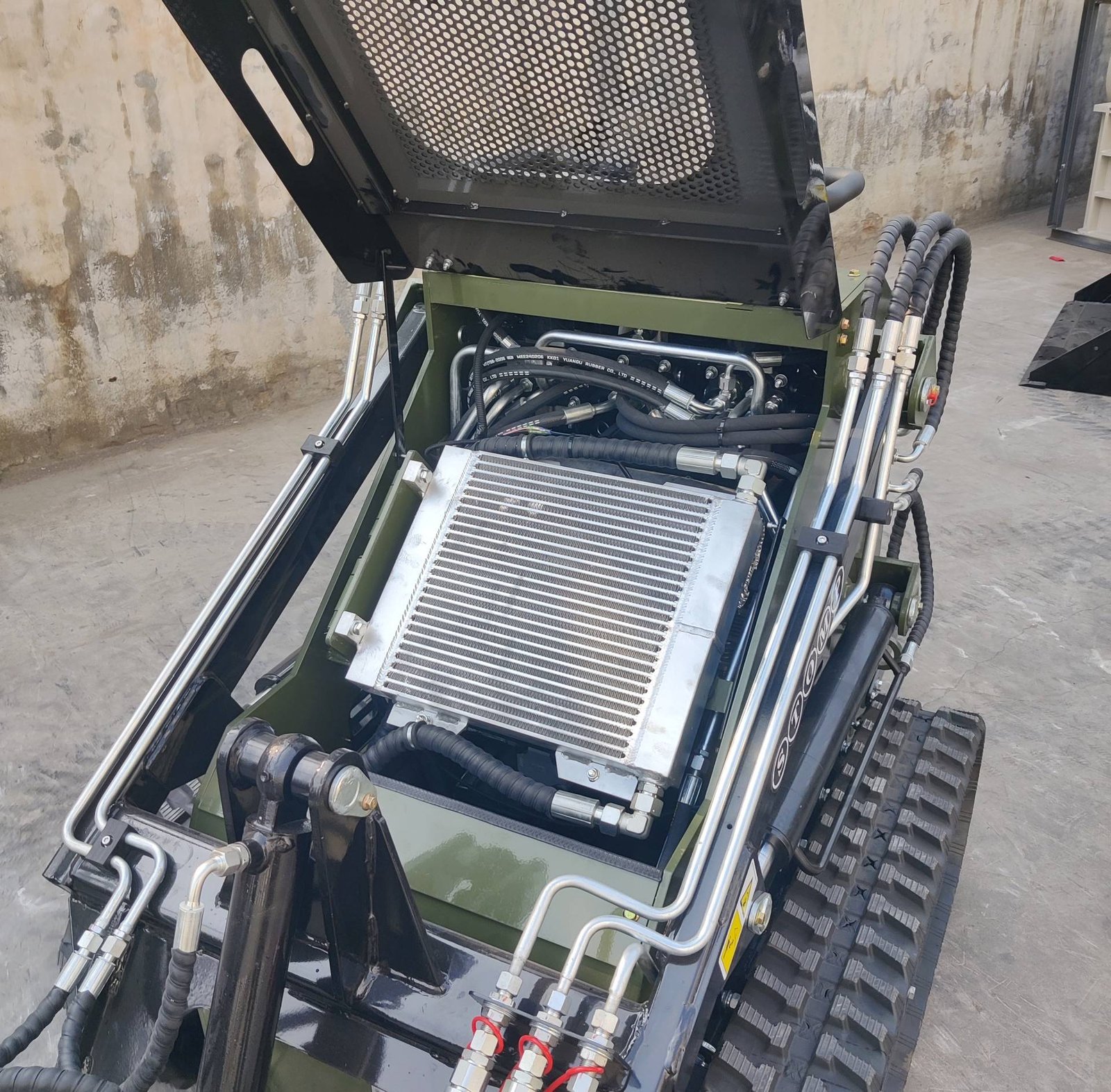 Hydraulic hose assembly with metal fittings mounted on a green industrial machine, featuring a perforated metal grate and exposed piping.