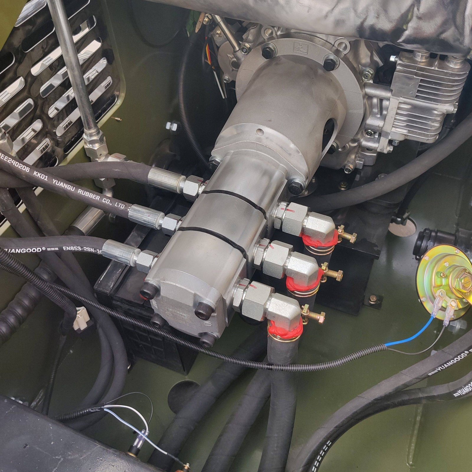 Industrial engine bay with metal pipes, hoses, and fittings; visible bolts and yellow-green equipment in background