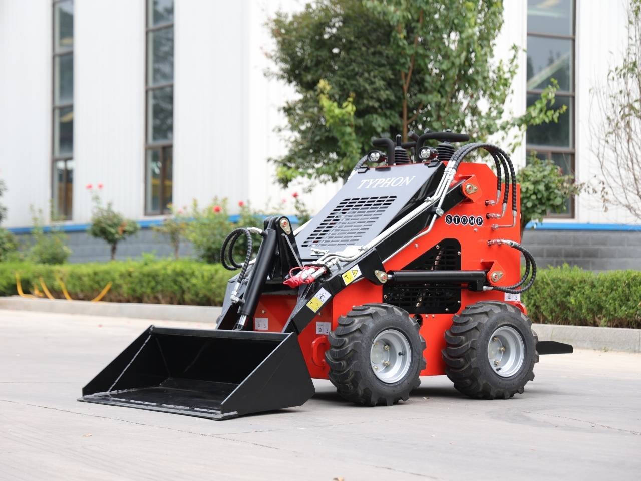 Red compact skid-steer loader with a front black bucket attachment on a paved surface near a landscaped area and building windows; machine labeled 'TYPHON'.