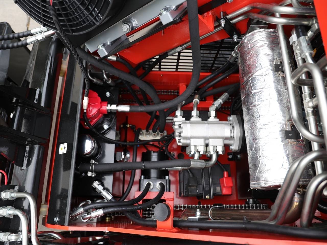 Close-up of a red chassis machinery interior showing a silver hydraulic pump connected to black hoses and metal tubing, with an insulated exhaust pipe on the right and various fittings around it.