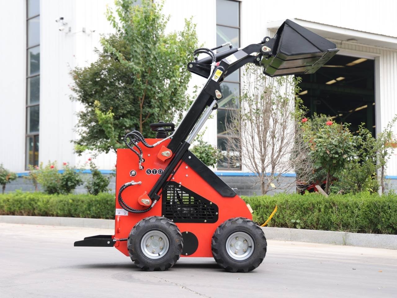 Red compact skid-steer loader with a raised black bucket, parked on a paved area near a white industrial building and greenery.