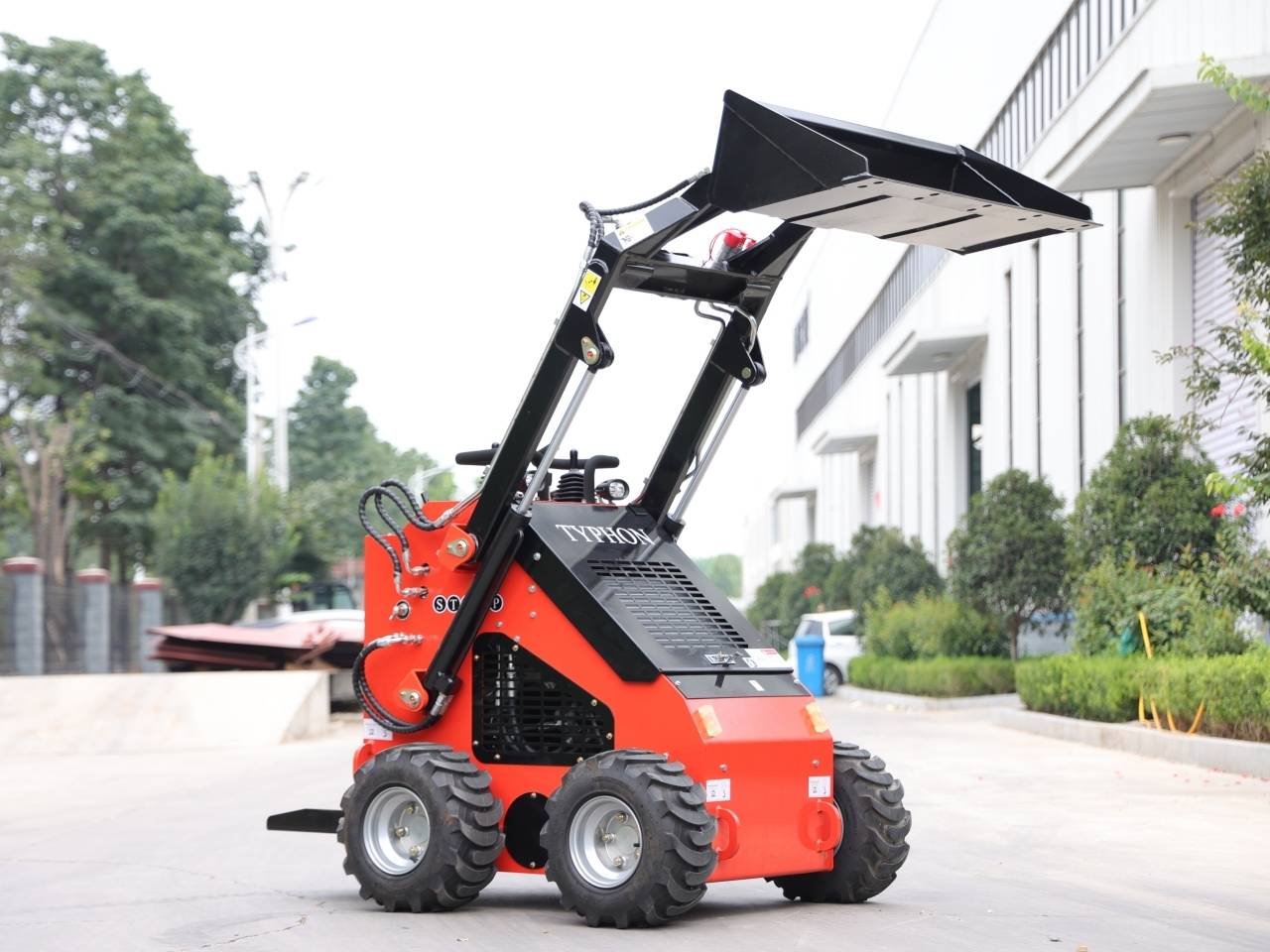 Red Typhoon skid-steer loader with a raised front bucket on a paved area near a building plot of greenery in the background