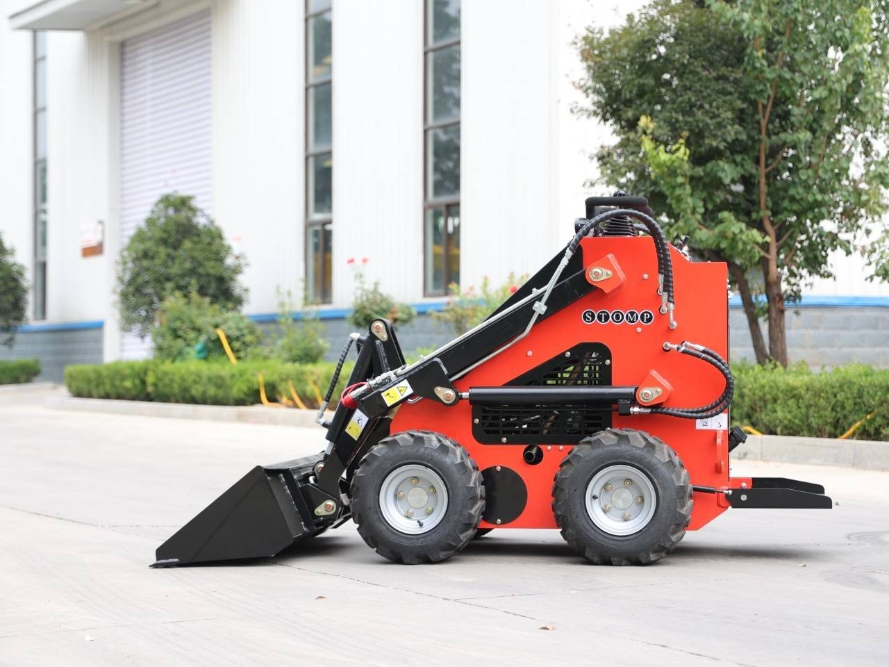 Red compact skid-steer loader with a front bucket parked on a paved area near an industrial building, ready for loading. Captures construction/landscaping equipment use.