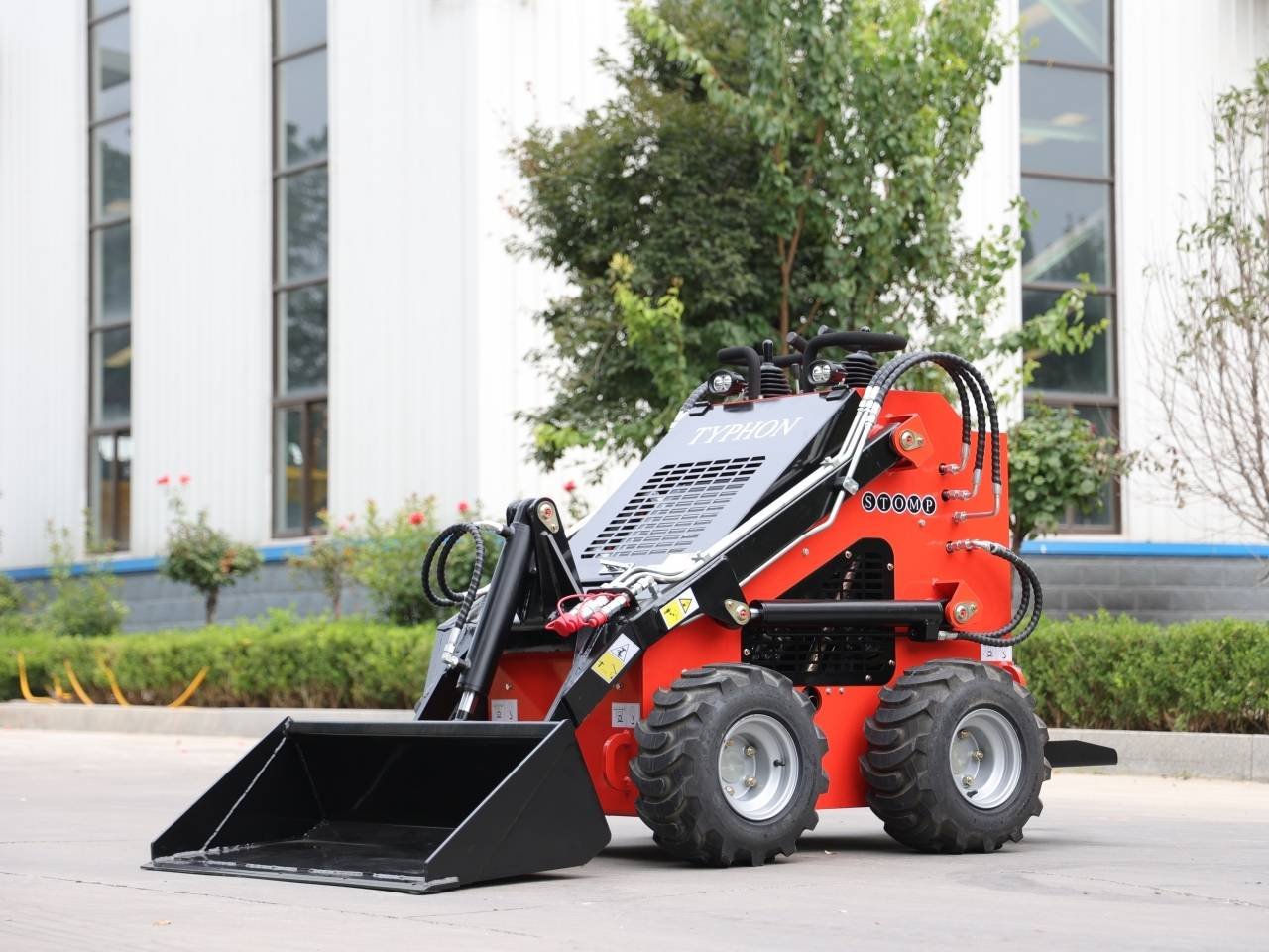 Red compact skid-steer loader with a front bucket parked on a paved area, outdoors near a building.