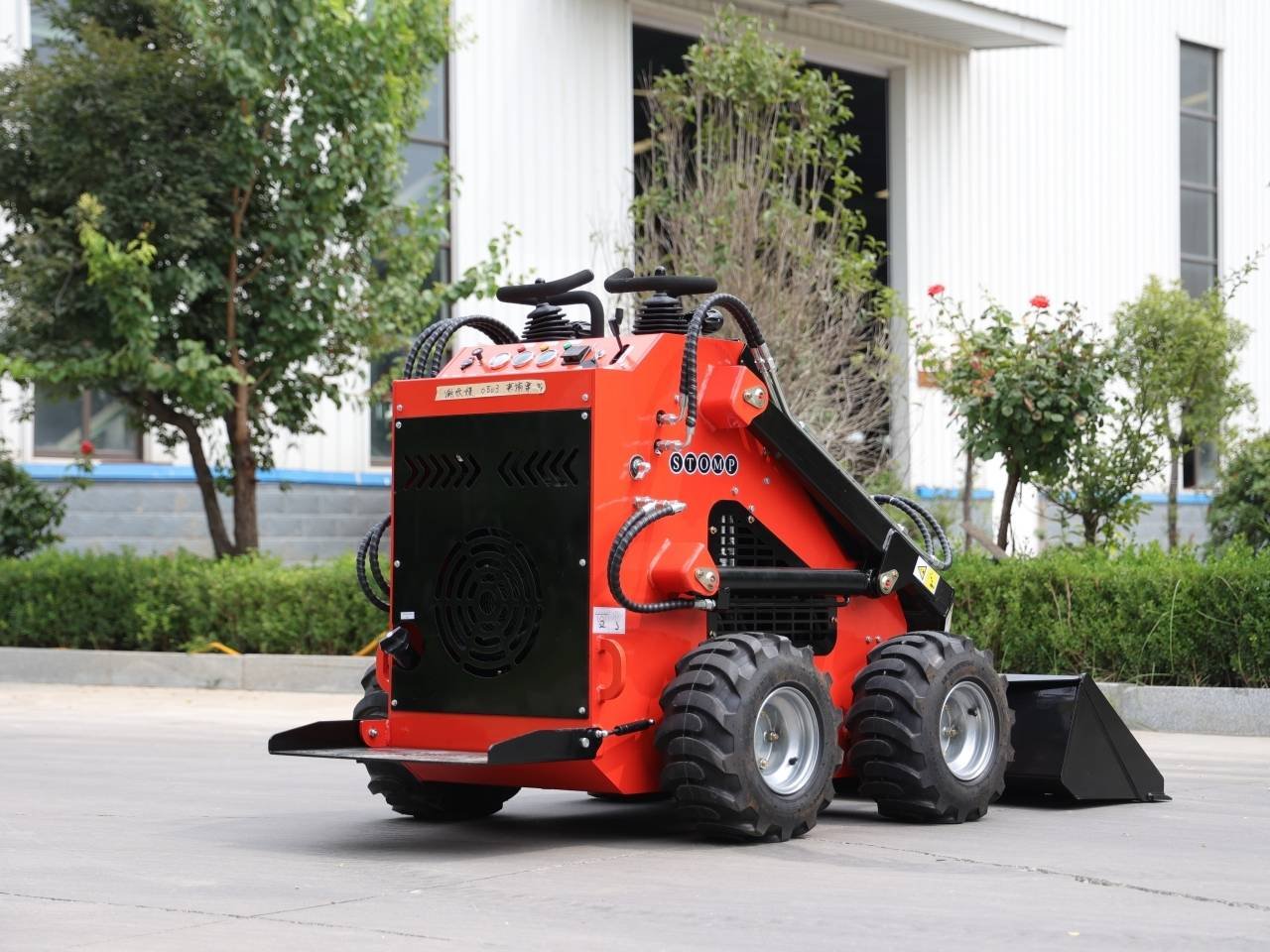 Red skid-steer loader with a front bucket parked on a paved area near a landscaped building garden.