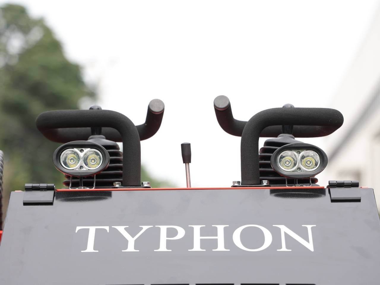 Front view of a dark gray machine labeled TYPHON with two dual-headlight pods and black padded handles above it, against a blurred outdoor background.