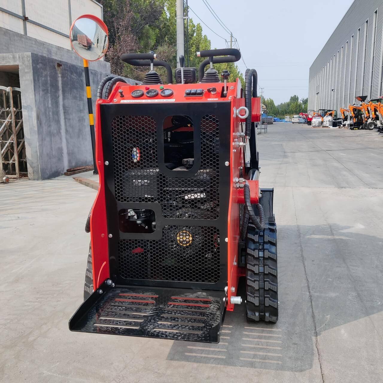 Red compact skid-steer loader with a black mesh engine guard, parked on a concrete yard with hydraulic arms and treaded tracks visible on the right side