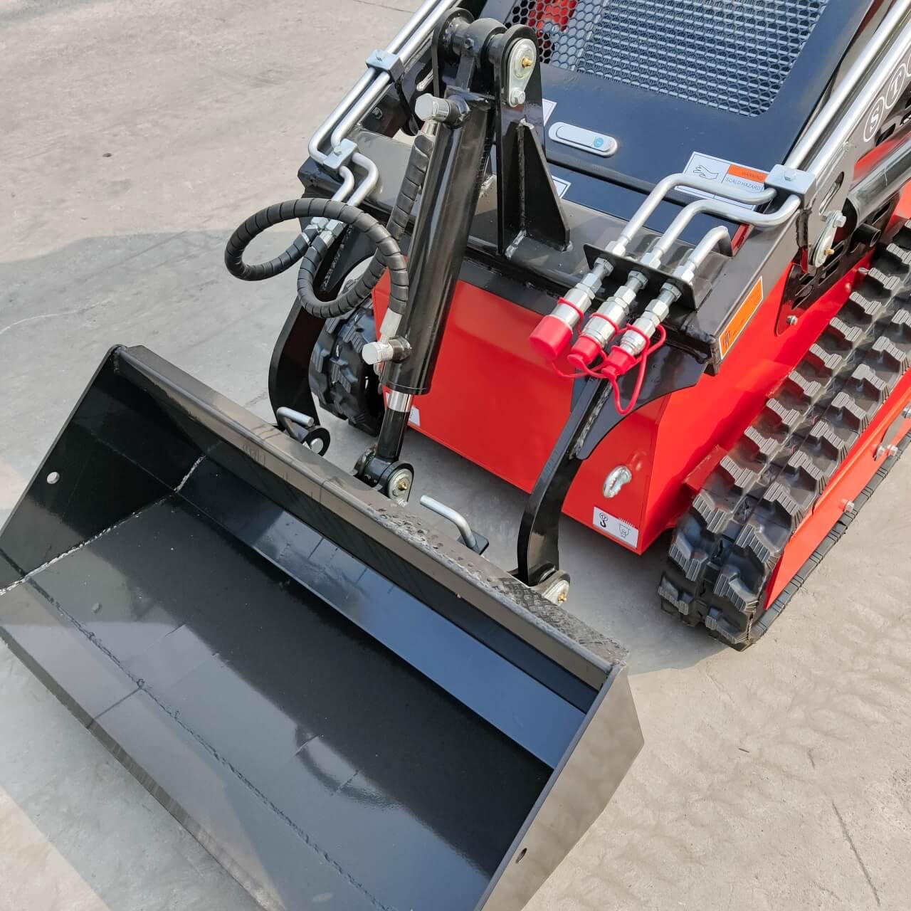 Red compact track loader with a large black front bucket and hydraulic arms on a concrete surface (close-up).