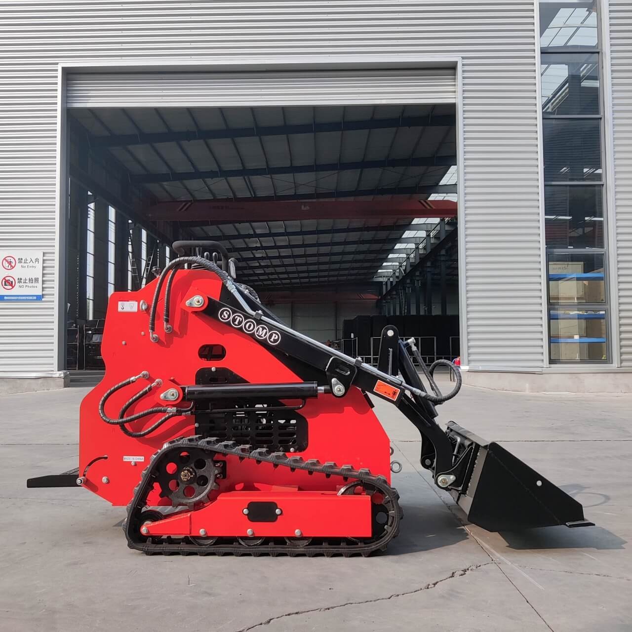 Red compact track loader with a front bucket attachment parked outside a metal industrial building. It has rubber tracks and hydraulic arms visible at the front edge of the loader caparus.
