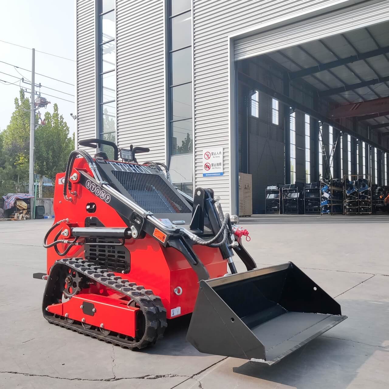 Red skid-steer loader with a front bucket parked outside a large metal warehouse, ready for use in construction.