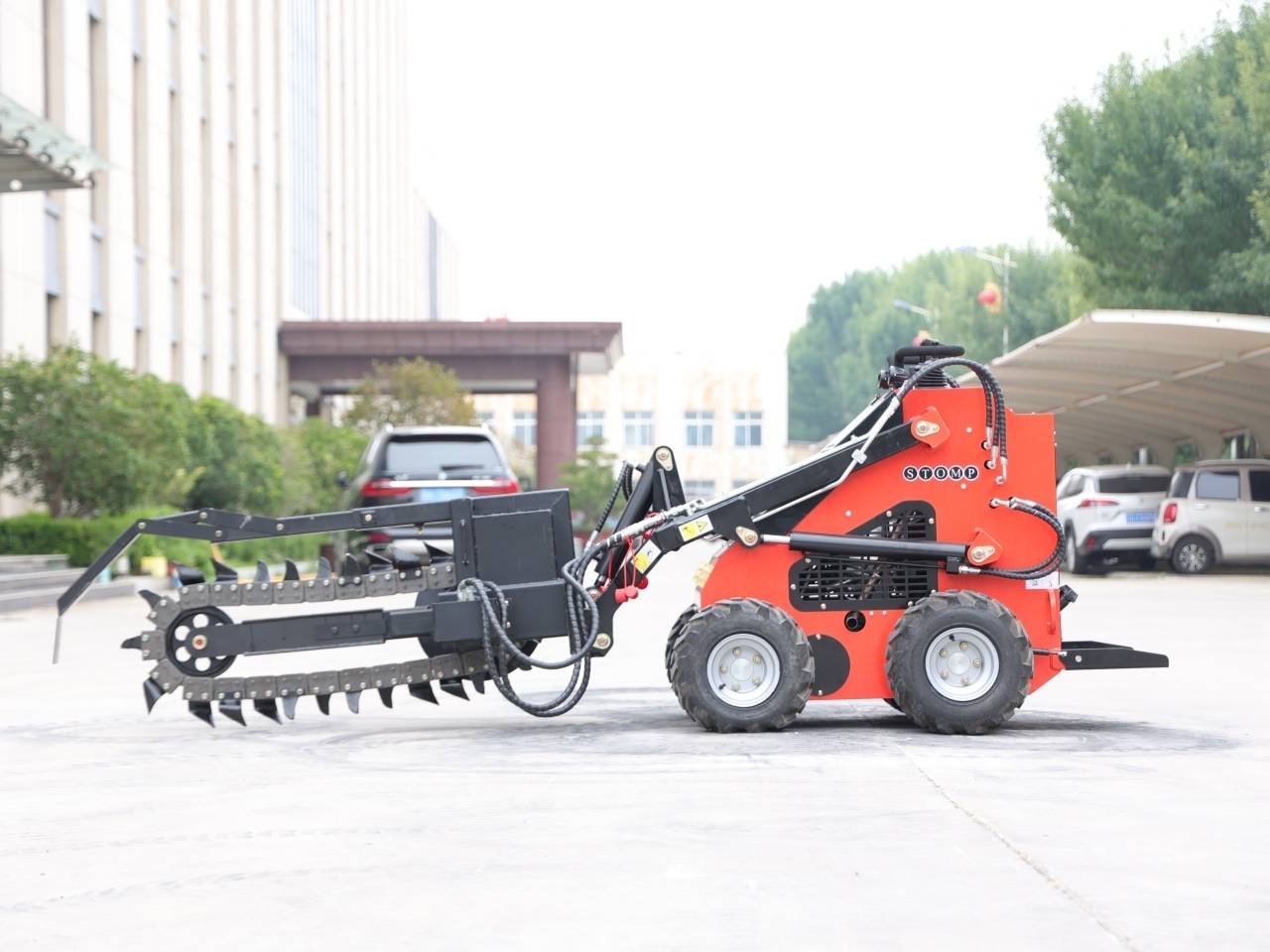 Red compact skid-steer loader with a large serrated rake attachment for land clearing, parked on a concrete lot outdoors in front of buildings and trees.