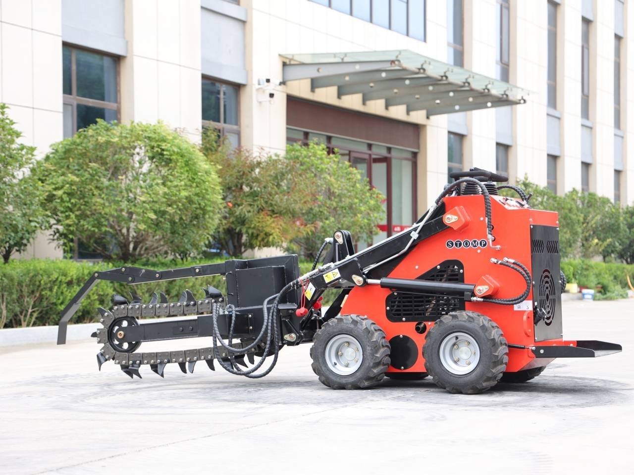 Red compact skid-steer loader with a hydraulic, toothed grapple attachment extended at the front, parked outside a modern building with greenery nearby.