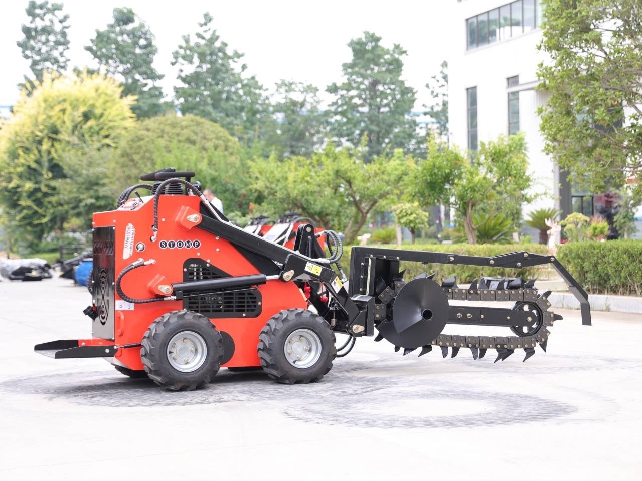 Red compact skid-steer loader with a large black hydraulic arm and a toothed trencher attachment extending to the right, parked on a paved area with trees and a building in the background